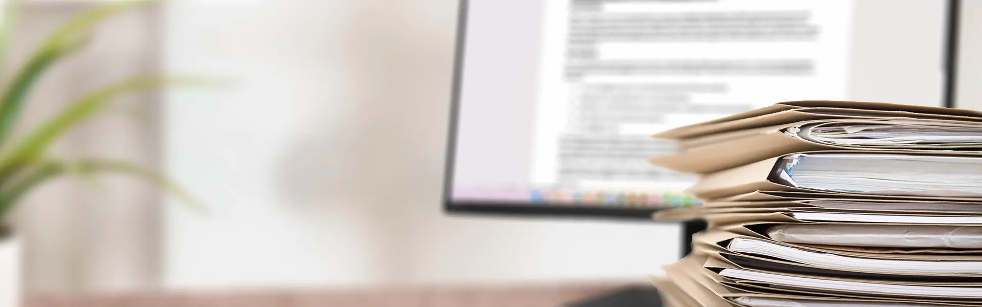 Stack of papers with a computer screen on a desk in the background.