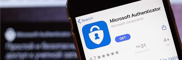 Downloading Microsoft Authenticator from the Apple App Store on an iPhone with a desktop computer in the background