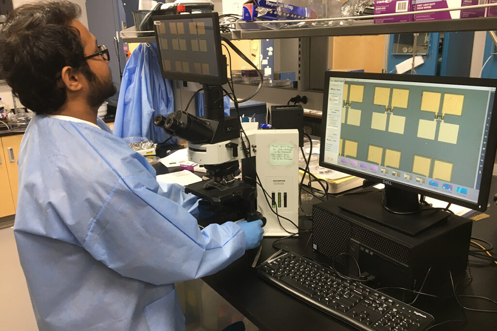 Man in a labcoat looking through a microscope via computer screen.